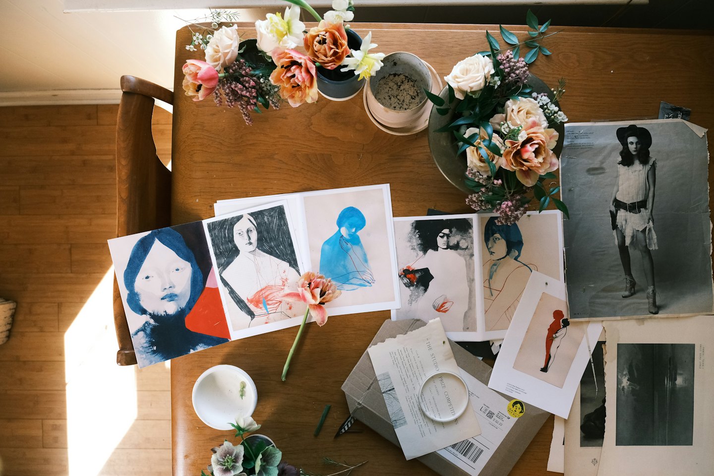 Wooden table with art prints, flowers, and a fashion photo.